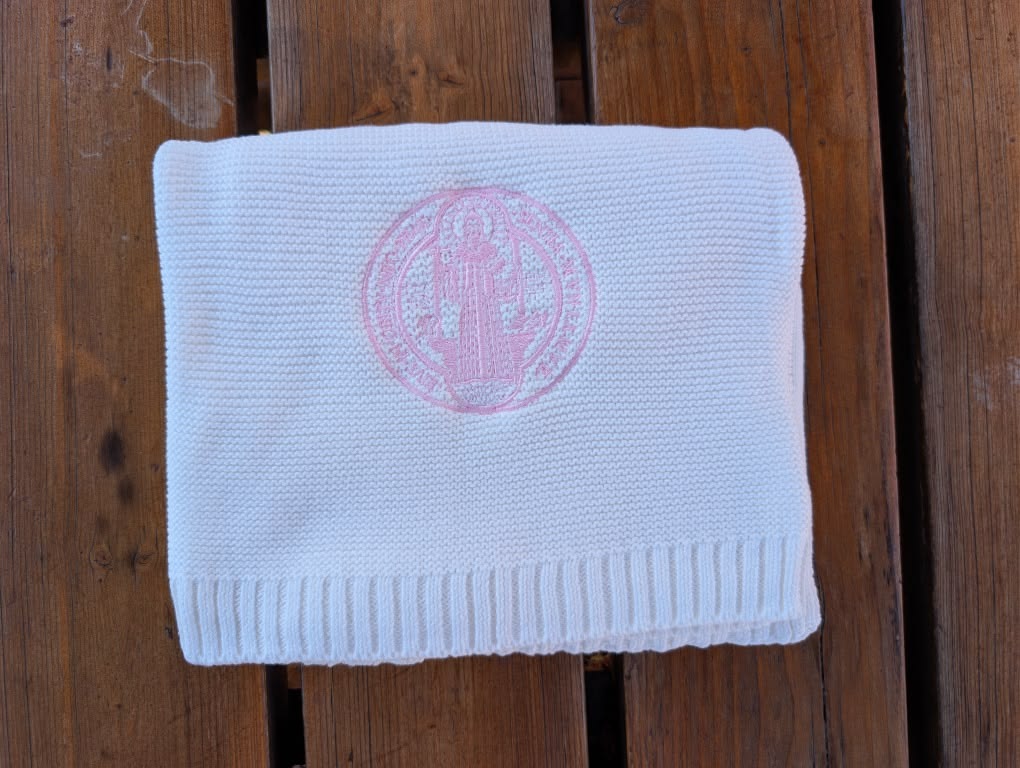 A folded white knit blanket with a pink St Benedict medal embroidered monogram, displayed on a brown wooden slatted surface.