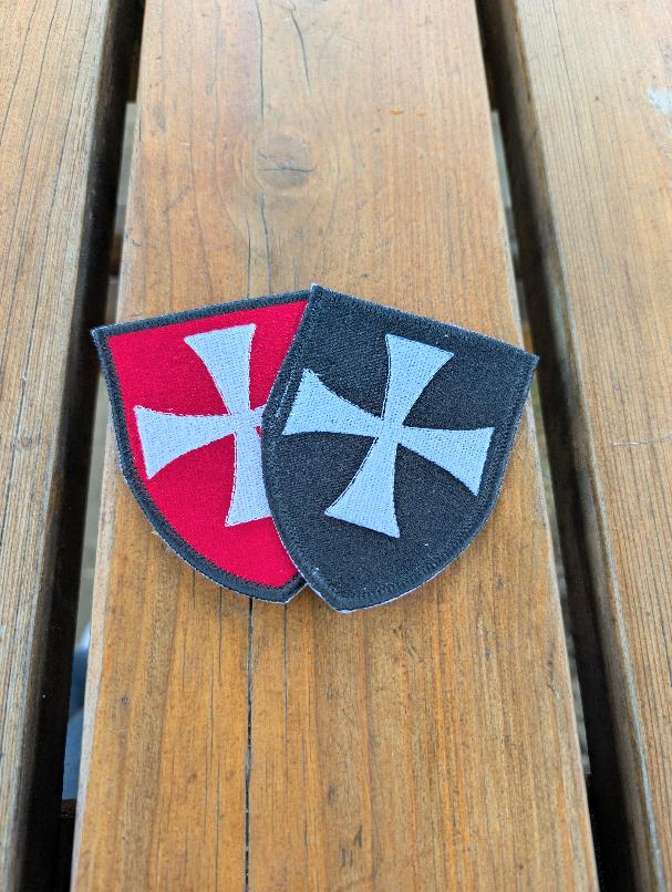 Two shield-shaped embroidered Knights Hospitaller badges arranged diagonally on a wooden background, with the red badge on the left and the black badge on the right.