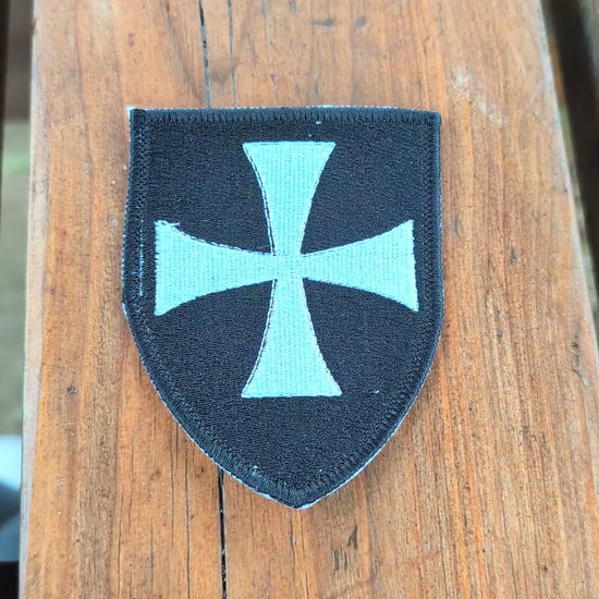 Video showing a black Knights Hospitaller embroidered badge on a wooden surface. The camera zooms in on the badge before it is turned over to show the backing, then flipped back to display the embroidered front again.