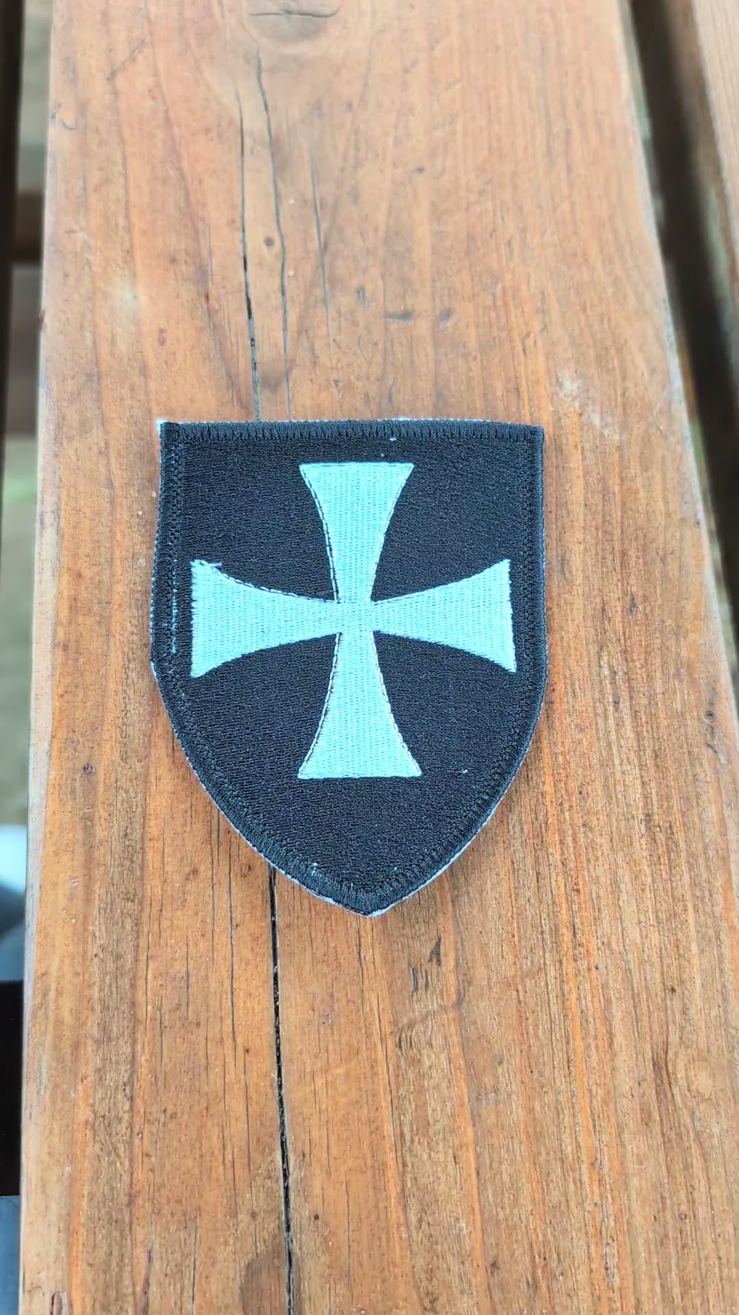 Video showing a black Knights Hospitaller embroidered badge on a wooden surface. The camera zooms in on the badge before it is turned over to show the backing, then flipped back to display the embroidered front again.