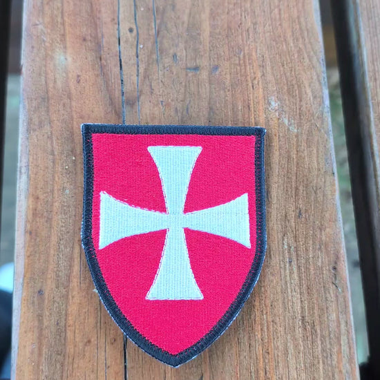 Video showing a red Knights Hospitaller embroidered badge on a wooden surface. The camera zooms in on the badge before it is turned over to show the backing, then flipped back to display the embroidered front again.