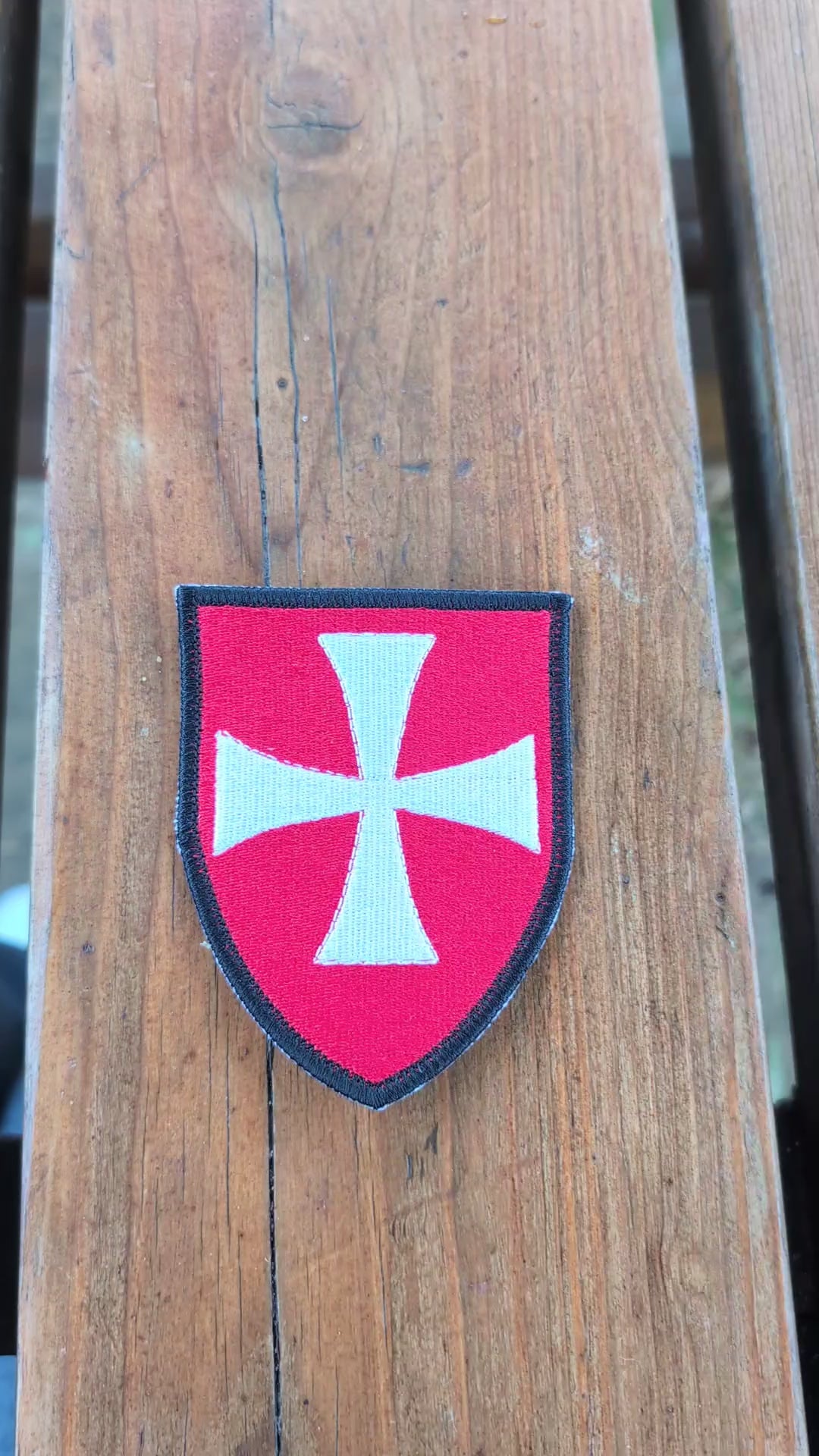 Video showing a red Knights Hospitaller embroidered badge on a wooden surface. The camera zooms in on the badge before it is turned over to show the backing, then flipped back to display the embroidered front again.