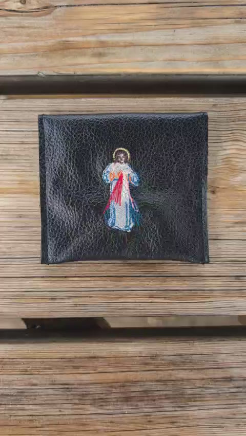 Video of a Divine Mercy leather rosary pouch on a wooden background, flipped to show the front and secure popper closure before returning to the original view