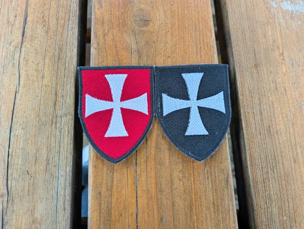 Two shield-shaped embroidered Knights Hospitaller badges displayed side by side on a wooden background, with the red badge on the left and the black badge on the right.